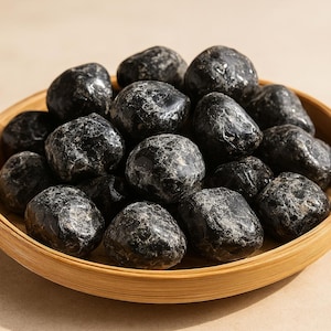 May include: A collection of polished black stones, possibly gemstones or decorative rocks, fills a round, light brown wooden bowl. The stones have a smooth, glossy surface with subtle variations in texture and color.