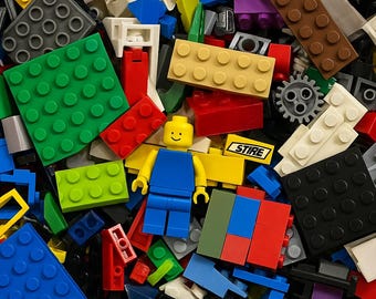 Puede incluir: Un colorido surtido de bloques de construcción de plástico y una minifigura amarilla con una cara sonriente. Los bloques son de varios colores, incluyendo rojo, verde, azul, amarillo y negro. Algunos bloques tienen clavijas y otros son lisos. La palabra "STIRE" es visible en un bloque amarillo.