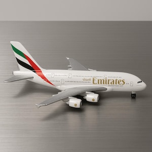 May include: A white Emirates airplane model with gray wings and engines. The tail features green, red, and black stripes. The Emirates logo is printed in gold on the side of the plane. The model has small details like windows and landing gear.