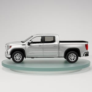 May include: A silver pickup truck model with black tires and rims. The truck has a four-door cab and a cargo bed. The model is displayed on a clear, round platform against a white background. The truck's headlights and taillights are visible.