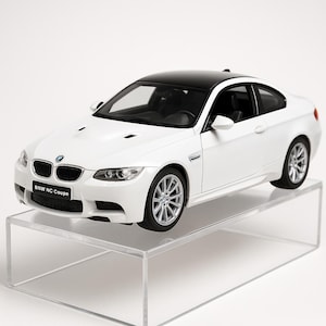 May include: A white BMW NC Coupe model car with a black roof and silver wheels. The car is displayed on a clear acrylic stand. The front of the car has the BMW logo and the text "BMW NC Coupe".
