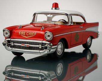 Op de afbeelding: Een rode en witte vintage speelgoedauto met de woorden "FIRE CHIEF" in gouden letters. De auto heeft chromen details, een rood licht op het dak en een reflecterend oppervlak eronder.