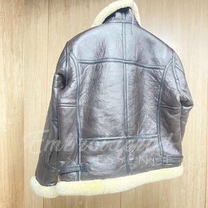 May include: A dark brown leather jacket featuring a thick cream-colored shearling collar, cuffs, and hem. The jacket has visible seam detailing, long sleeves, and adjustable side straps at the waist. The surface has a textured, glossy finish.
