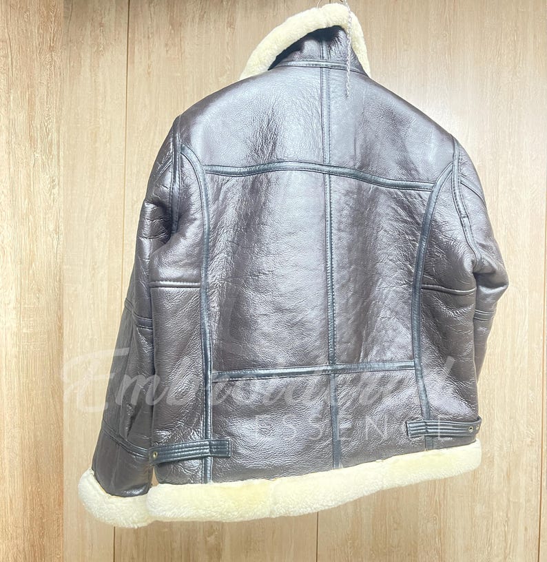May include: A dark brown leather jacket featuring a thick cream-colored shearling collar, cuffs, and hem. The jacket has visible seam detailing, long sleeves, and adjustable side straps at the waist. The surface has a textured, glossy finish.
