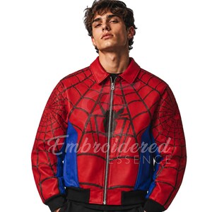 K&ouml;nnte beinhalten: Rote und blaue Jacke im Spider-Man-Design mit schwarzem Spinnennetz-Muster und schwarzem Spinnen-Emblem. Die Jacke hat einen Rei&szlig;verschluss, einen schwarzen Kragen und blaue Seitenteile. Der Text "Embroidered Essence" ist sichtbar.