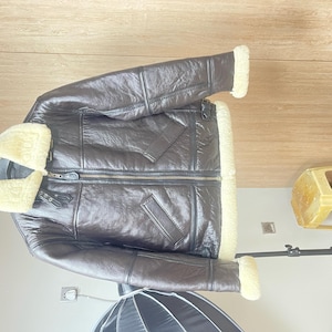 May include: A dark brown leather jacket featuring a thick cream-colored shearling lining, a wide fold-over collar, a central front zipper closure, two slanted side pockets, and adjustable buckle straps at the collar and waist for a secure fit.