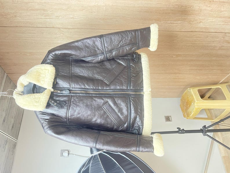 May include: A dark brown leather jacket featuring a thick cream-colored shearling lining, a wide fold-over collar, a central front zipper closure, two slanted side pockets, and adjustable buckle straps at the collar and waist for a secure fit.