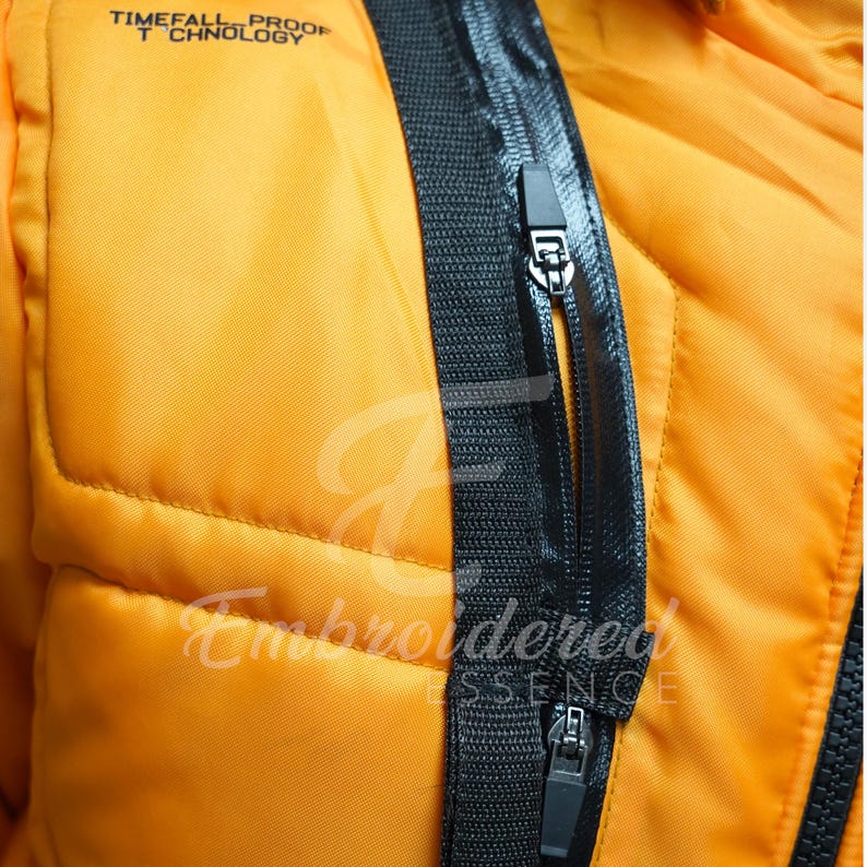 May include: Close-up of an orange jacket with black zippers and trim. The jacket has a quilted design and the text "TIMEFALL PROOF TECHNOLOGY" is visible at the top. The words "Embroidered Essence" are also visible.