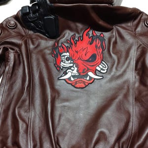 May include: A brown leather jacket featuring a large embroidered red and black oni mask graphic on the back. The collar displays a black patch with the white text SAMURAI. A black holster-like attachment is secured to the upper left shoulder area.