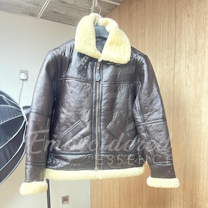 May include: A dark brown jacket with a textured finish, featuring a cream-colored faux shearling collar and cuffs. The jacket includes a central metal zipper, two slanted front pockets, and a buckled strap detail at the neck for closure.