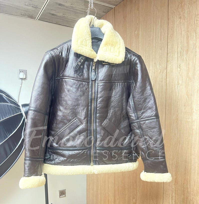 May include: A dark brown jacket with a textured finish, featuring a cream-colored faux shearling collar and cuffs. The jacket includes a central metal zipper, two slanted front pockets, and a buckled strap detail at the neck for closure.