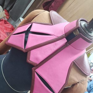 May include: A structured chest piece featuring bright pink panels with dark brown trim and accents. The design includes geometric cutouts revealing a black base layer, a high collar, and shoulder guards, all displayed on a mannequin torso.
