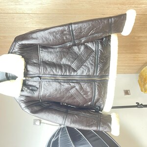May include: A dark brown leather jacket featuring a thick cream-colored shearling lining, a wide fold-over collar, long sleeves with shearling cuffs, a central metal zipper closure, and two slanted front pockets with leather trim detailing.