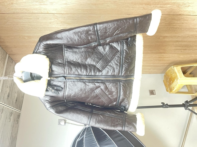 May include: A dark brown leather jacket featuring a thick cream-colored shearling lining, a wide fold-over collar, long sleeves with shearling cuffs, a central metal zipper closure, and two slanted front pockets with leather trim detailing.
