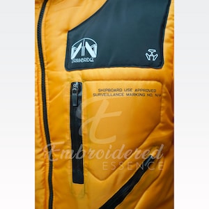 May include: Close-up of a yellow quilted jacket with black accents. Features a black pocket with the text "SHIPBOARD USE APPROVED SURVEILLANCE MARKING NO, N/A". The jacket has a black zipper and the text "DRAWBRIDGE". The brand name "Embroidered ESSENCE" is also visible.