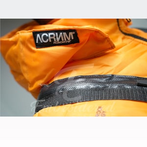 May include: Close-up of an orange jacket with a black zipper and the brand name "ACRONYM" printed on a black label. The jacket appears to be made of a durable, possibly waterproof material.
