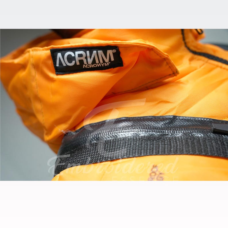 May include: Close-up of an orange jacket with a black zipper and the brand name "ACRONYM" printed on a black label. The jacket appears to be made of a durable, possibly waterproof material.