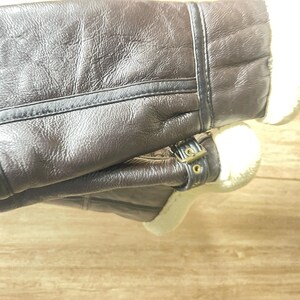 May include: A pair of dark brown leather boots featuring a textured surface, visible stitched seams, and a soft white faux fur lining. The boots include a decorative strap with a small metallic buckle detail positioned near the ankle area.