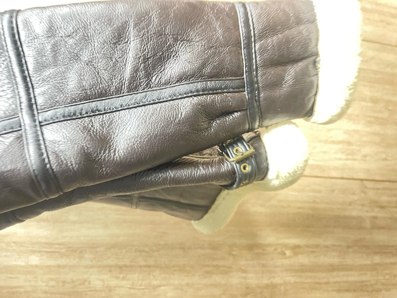 May include: A pair of dark brown leather boots featuring a textured surface, visible stitched seams, and a soft white faux fur lining. The boots include a decorative strap with a small metallic buckle detail positioned near the ankle area.