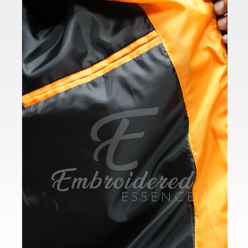May include: Close-up of the inside of a jacket. The interior is black with an orange trim and accents. The jacket has a pocket on the left side. The text "Embroidered Essence" is printed on the jacket.