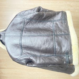 May include: A dark brown leather jacket featuring a thick cream-colored shearling lining along the collar and front opening. The jacket has visible seam detailing across the back and sides, with adjustable leather straps at the lower waist area.