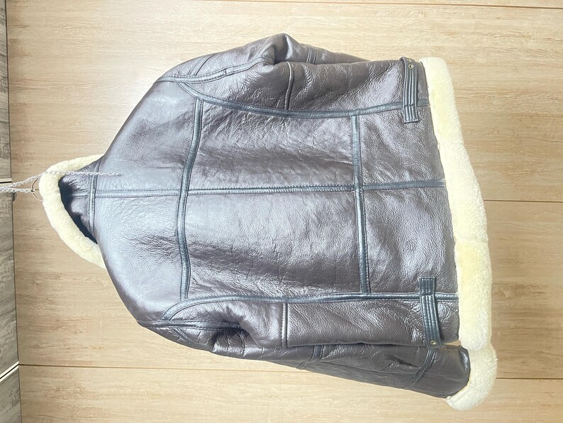 May include: A dark brown leather jacket featuring a thick cream-colored shearling lining along the collar and front opening. The jacket has visible seam detailing across the back and sides, with adjustable leather straps at the lower waist area.
