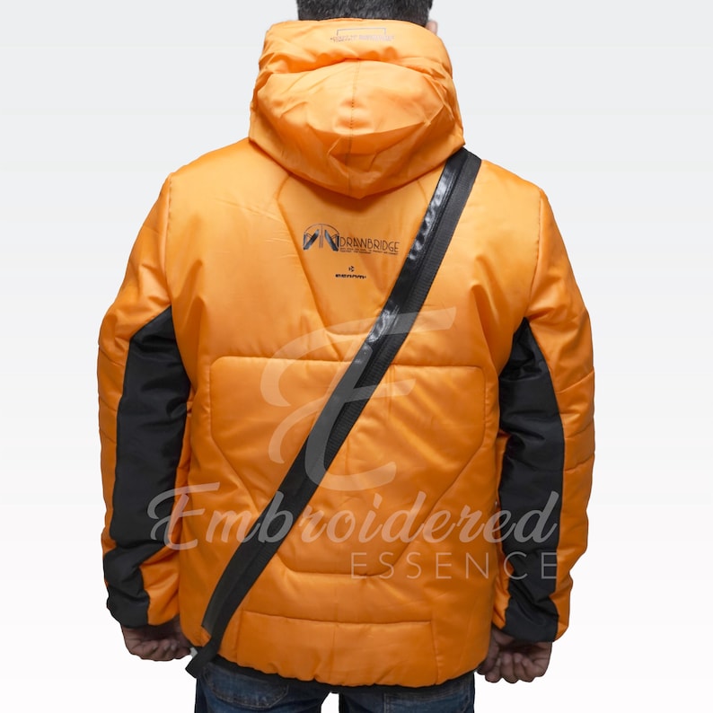May include: Orange puffer jacket with black accents on the sleeves and a diagonal black strap across the back. The jacket has a hood and features the text "Embroidered Essence" and a logo on the upper back.