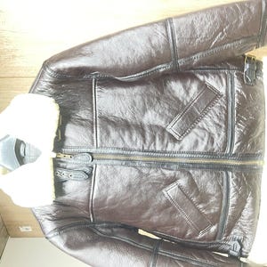 May include: A dark brown jacket with a glossy finish, featuring a thick white faux shearling collar and interior lining. The jacket includes a central brass-toned zipper, two slanted front pockets, and black leather-like trim along the seams.