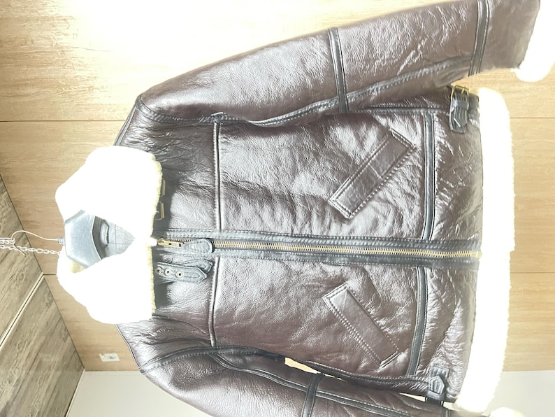 May include: A dark brown jacket with a glossy finish, featuring a thick white faux shearling collar and interior lining. The jacket includes a central brass-toned zipper, two slanted front pockets, and black leather-like trim along the seams.