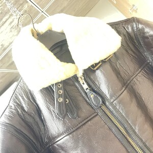 May include: A dark brown leather jacket featuring a thick, cream-colored faux fur collar. The jacket includes a central metal zipper, two decorative leather tabs with silver-toned metal eyelets near the neckline, and visible seam detailing.