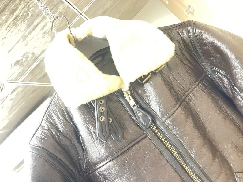 May include: A dark brown leather jacket featuring a thick, cream-colored faux fur collar. The jacket includes a central metal zipper, two decorative leather tabs with silver-toned metal eyelets near the neckline, and visible seam detailing.
