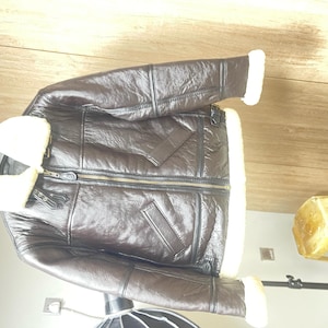 May include: A dark brown leather jacket featuring a thick white shearling collar and matching white shearling trim at the sleeve cuffs and hem. The jacket includes a central front zipper closure, two slanted side pockets, and adjustable buckle straps.