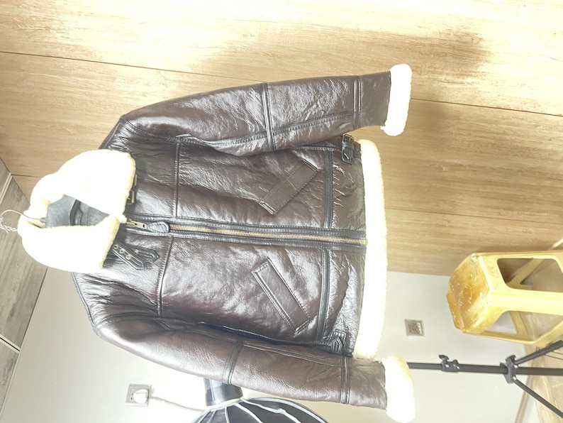 May include: A dark brown leather jacket featuring a thick white shearling collar and matching white shearling trim at the sleeve cuffs and hem. The jacket includes a central front zipper closure, two slanted side pockets, and adjustable buckle straps.