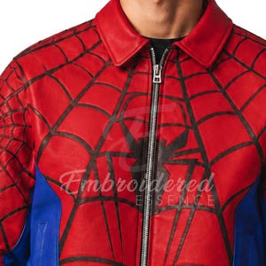 K&ouml;nnte beinhalten: Rote Lederjacke mit schwarzem Spinnennetz-Design und gro&szlig;em Spinnen-Emblem auf der Brust. Die Jacke hat einen Rei&szlig;verschluss und blaue Seitenteile. Der Text "Embroidered Essence" ist sichtbar.