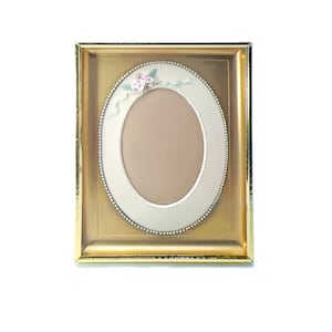 May include: A gold-colored oval picture frame with a white mat and a delicate floral design. The frame has a beaded border and a raised edge.