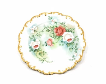 Peut inclure: Une assiette en porcelaine blanche avec un bord doré et un motif peint à la main de roses roses et blanches avec des feuilles vertes.