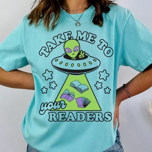 May include: A light blue t-shirt featuring a graphic of a green alien in a flying saucer beaming down books. The text reads "TAKE ME TO your READERS" in black lettering. The design includes small star shapes surrounding the central alien illustration.
