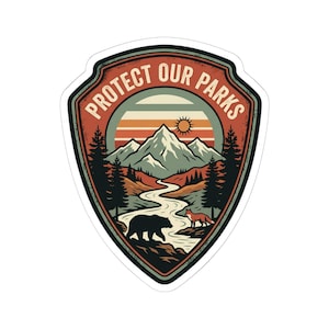 May include: A shield-shaped sticker featuring a landscape illustration with the text PROTECT OUR PARKS. The design includes snow-capped mountains, a winding river, pine trees, a bear, and a fox under a sunset sky with orange and teal tones.