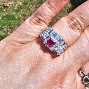 Op de afbeelding: Een zilveren ring met een vierkante, rode edelsteen in het midden, omringd door kleinere, heldere stenen. De ring zit om een vinger, met een tweede ring met een groene edelsteen op de achtergrond.