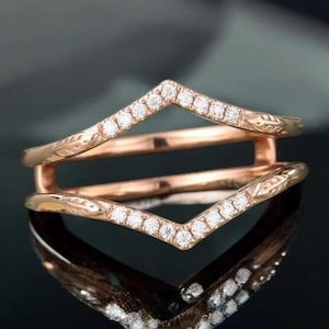 Op de afbeelding: Roségouden ring met een V-vormig ontwerp, versierd met kleine, sprankelende diamanten. De ring heeft een gegraveerd bloemmotief op de band. De ring wordt weerspiegeld op een donker, glanzend oppervlak.