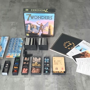 Puede incluir: Juego de mesa "7 Wonders" con una caja que muestra una figura con una antorcha. Incluye componentes del juego como cartas, fichas y reglas. El juego está sobre una superficie gris.