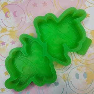 May include: Two bright green silicone molds in the shape of a cartoon character. The molds are on a colorful background with smiley faces and stars. The molds are likely for crafting or baking.