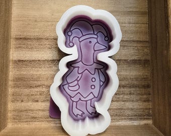 May include: A white silicone mold featuring a whimsical duck character dressed in a jester's outfit. The duck has a pointed hat with bells and a ruffled collar. The mold is set against a wooden background.