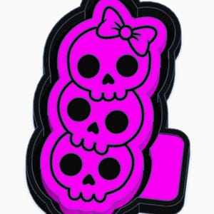 May include: A stacked design featuring three pink skulls with black eye sockets and a bow on top. The skulls are outlined in black, creating a 3D effect. The design has a rectangular base.