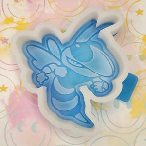 May include: A light blue silicone mold in the shape of a cartoon bee. The bee has a smiling face and outstretched arms. The mold is surrounded by a white border and sits on a colorful background with smiley faces and stars.