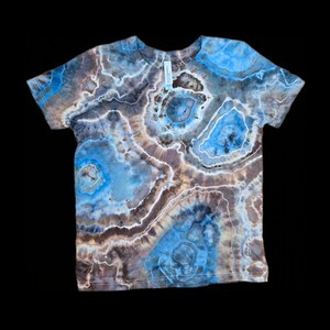 以下が含まれることがあります： 青、茶色、白の色合いのタイダイ柄の半袖Tシャツ。デザインは同心円と不規則な形状が特徴で、ユニークで抽象的な外観を作り出しています。シャツは柔らかい素材で作られています。