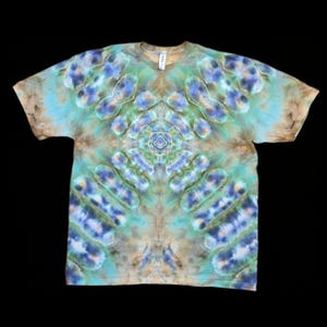 May include: A tie-dye t-shirt with a unique pattern in shades of blue, green, and brown. The design is symmetrical, with repeating shapes and a central floral-like motif. The shirt has a crew neck and short sleeves, and is displayed against a black background.