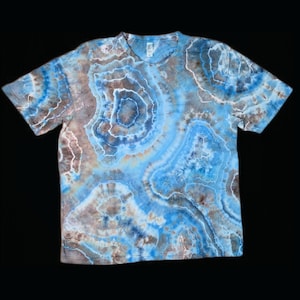 May include: A short-sleeved t-shirt with a tie-dye pattern in shades of blue, brown, and white. The design features concentric circular patterns, creating a marbled effect. The shirt has a crew neck and is made of a soft material.