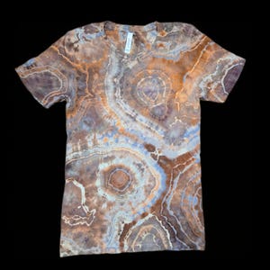 Pode incluir: Uma t-shirt de manga curta com um padrão tie-dye único. A t-shirt apresenta uma mistura de tons castanhos, laranja e azuis, criando um efeito marmoreado. O design inclui círculos concêntricos e linhas fluidas, dando-lhe um aspeto natural e orgânico. A t-shirt é feita de um material macio.