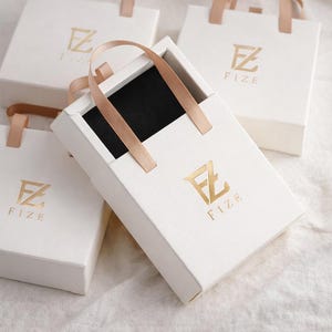 May include: White gift boxes with gold-colored logos and tan ribbon handles. The boxes are rectangular with a square base. The logo includes the letters "F" and "Z" with the word "FIZE" below. One box is open, revealing a black interior.